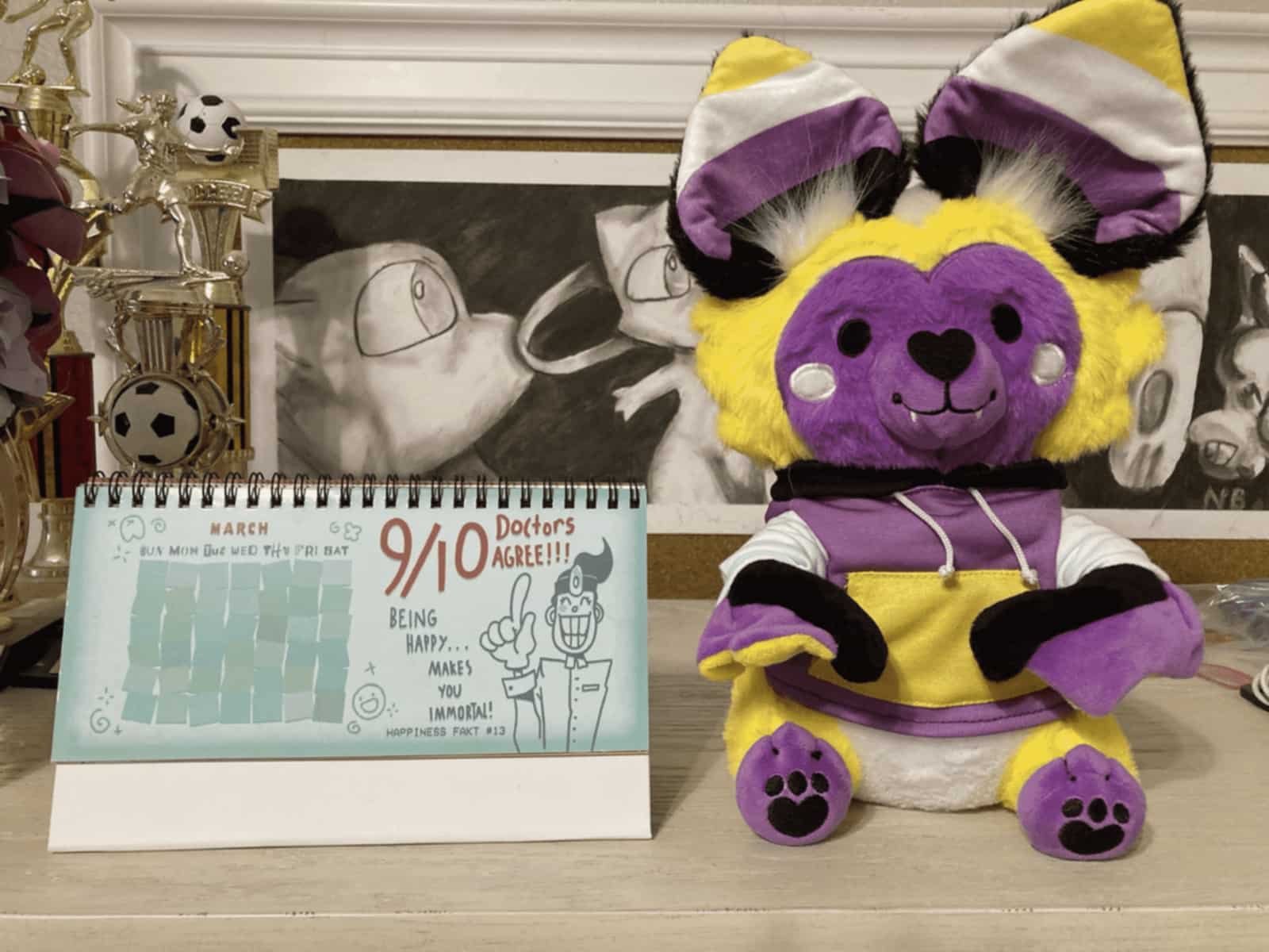A photo of the top of a piece of white furniture. On top of it, there is a Smile For Me calender, turned to March, and a nonbinary-colored and -themed bat plushie.